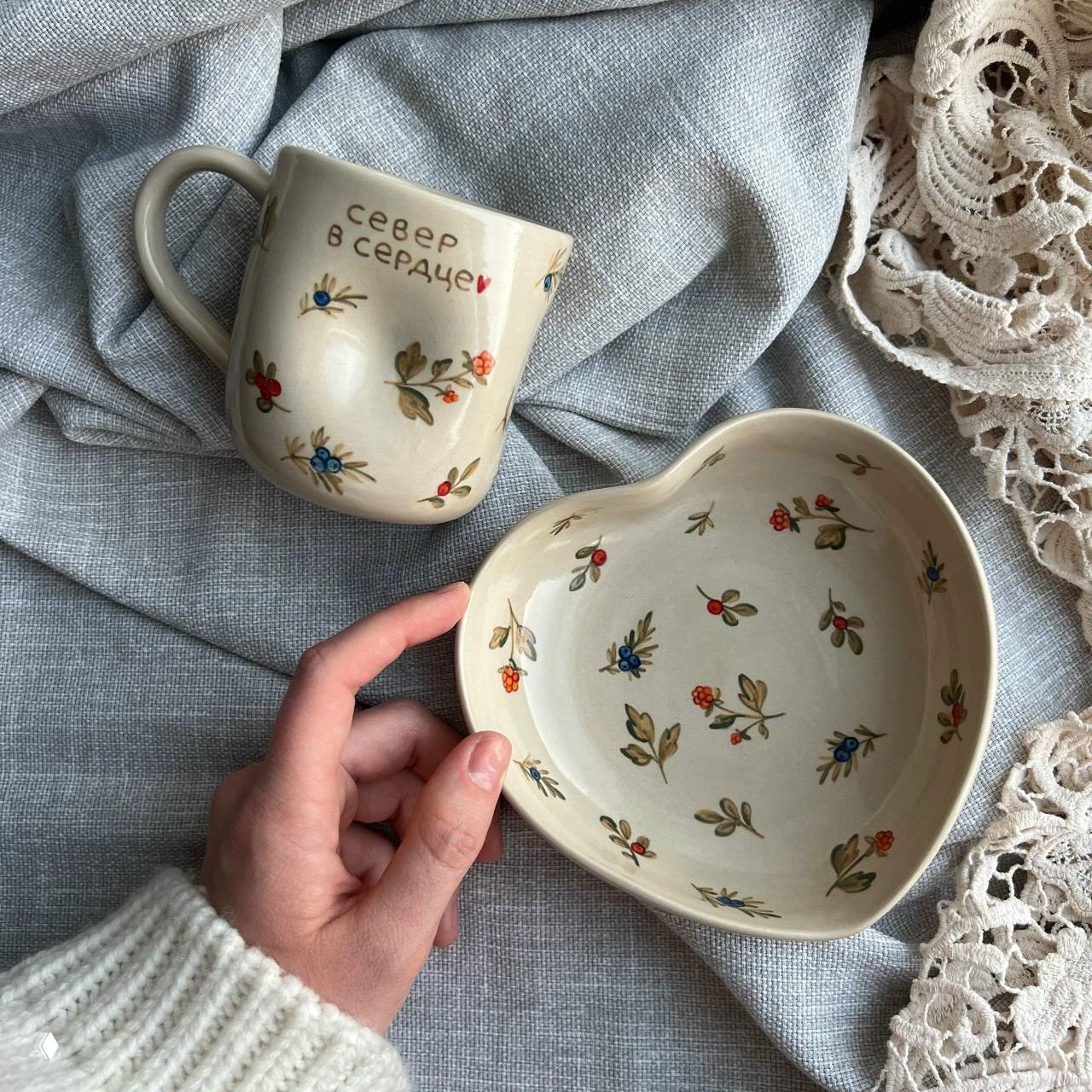 Ручная керамическая кружка с надписью «север в сердце» и тарелка в форме сердца с мелким растительным орнаментом на светлом текстильном фоне.