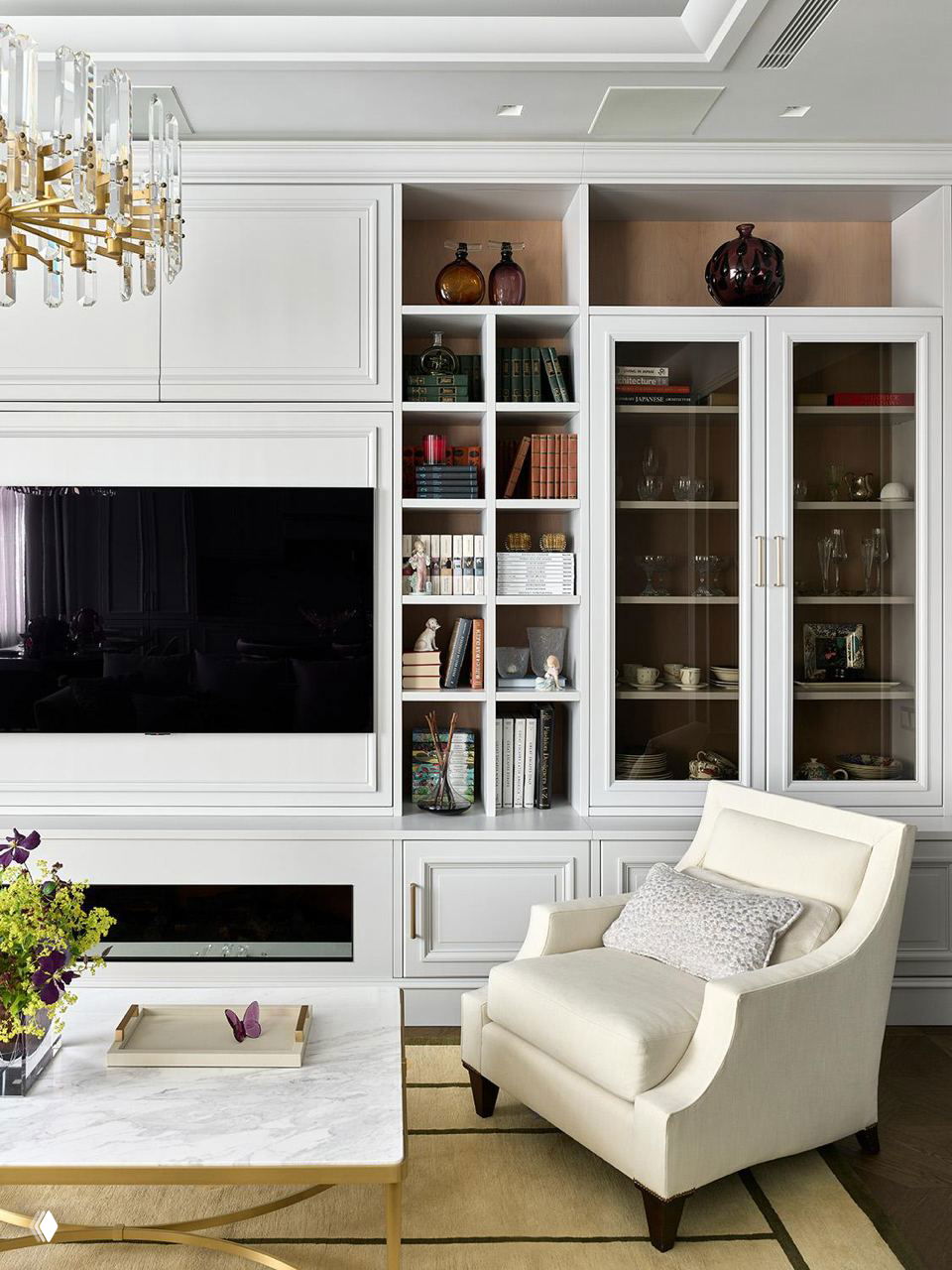 Гостиная квартиры 200 м² в центре Москвы: белая встроенная стена с телевизором, кремовое кресло, мраморный столик и изящная латунная люстра над зоной.