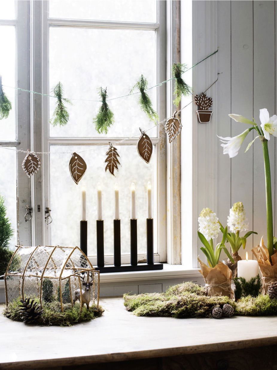Окно с подвешенными гирляндами из имбирных пряников и хвои; на подоконнике декоративные мхи, подсвечник и композиции в скандинавском стиле.