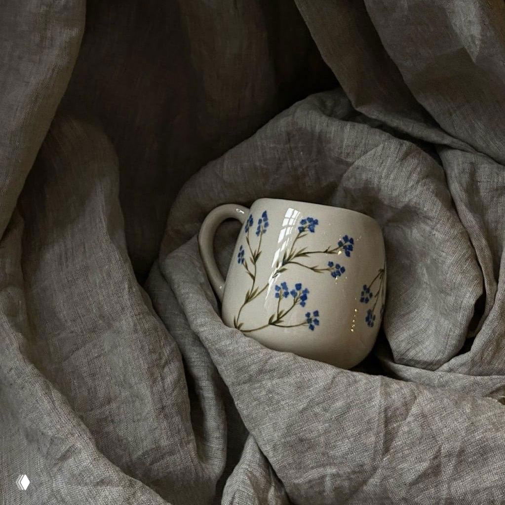 Керамическая кружка с мелким синим цветочным орнаментом, размещённая на складках серой льняной ткани — предметная фотография с уютной атмосферой фестиваля.