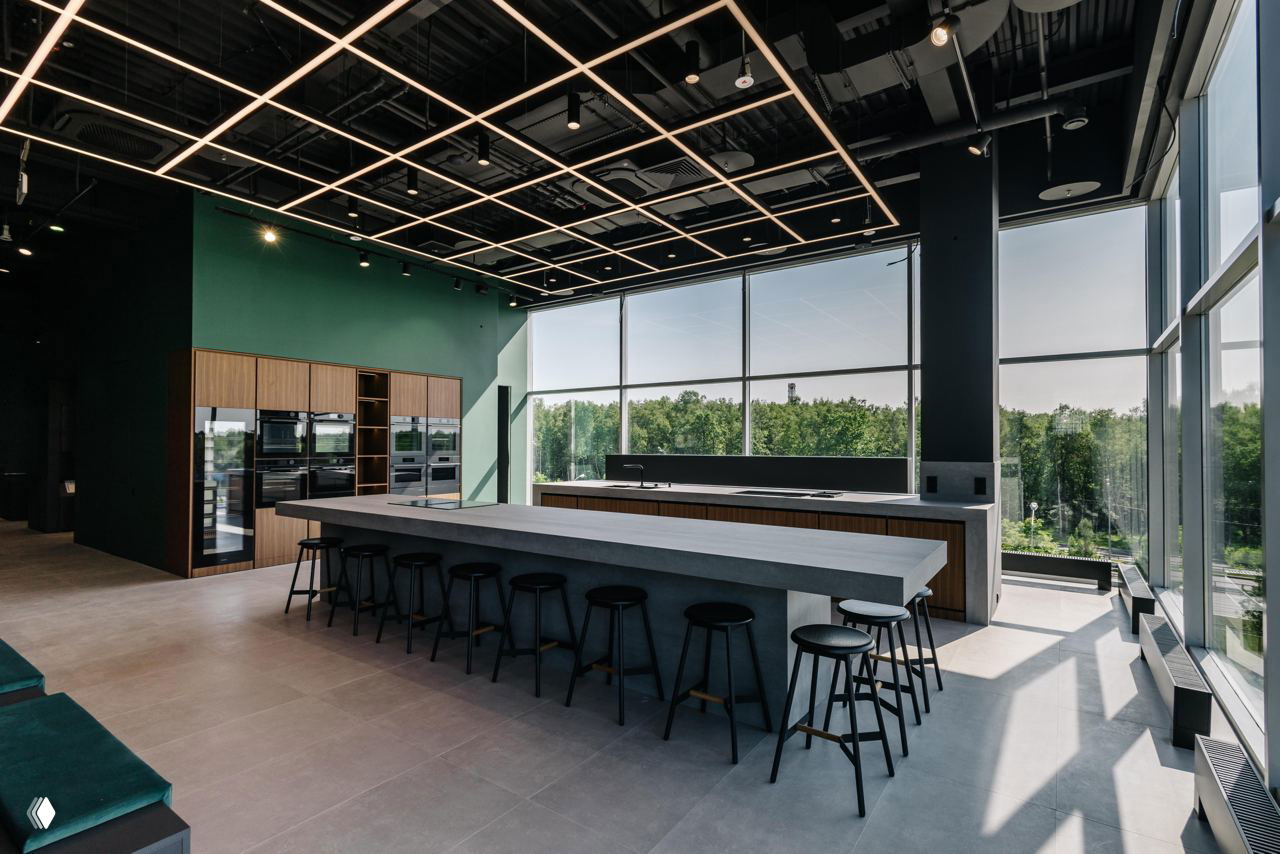 Большой стол для мастер-классов и барная зона в шоуруме High Level: длинная поверхность, табуреты, современная световая сетка и вид на зелёный пояс за окнами.