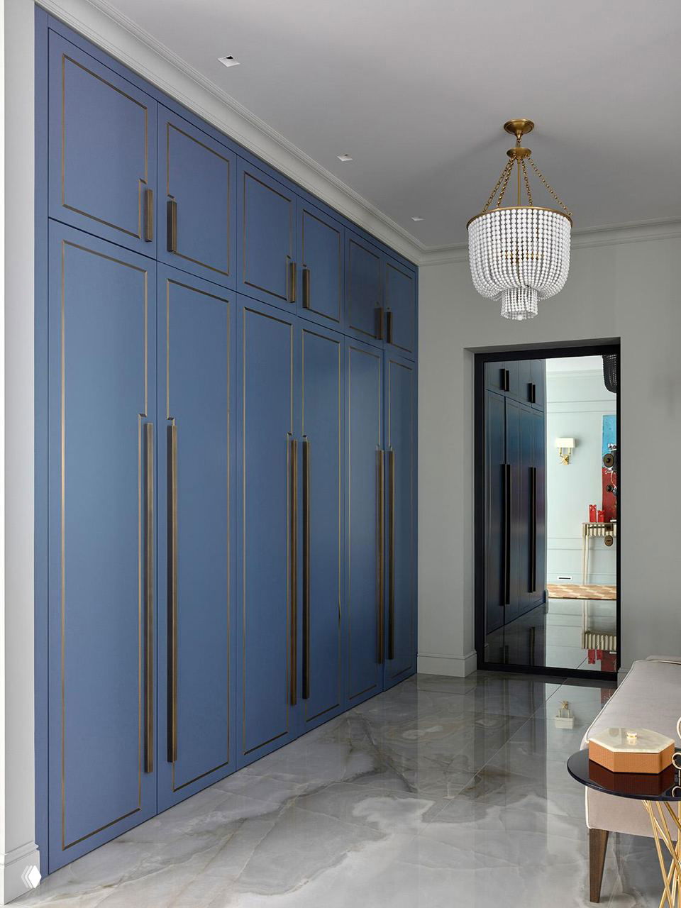 Коридор с высокими насыщенно-синими шкафами, длинными латунными ручками, глянцевым мраморным полом и подвесной хрустальной люстрой в современном интерьере.