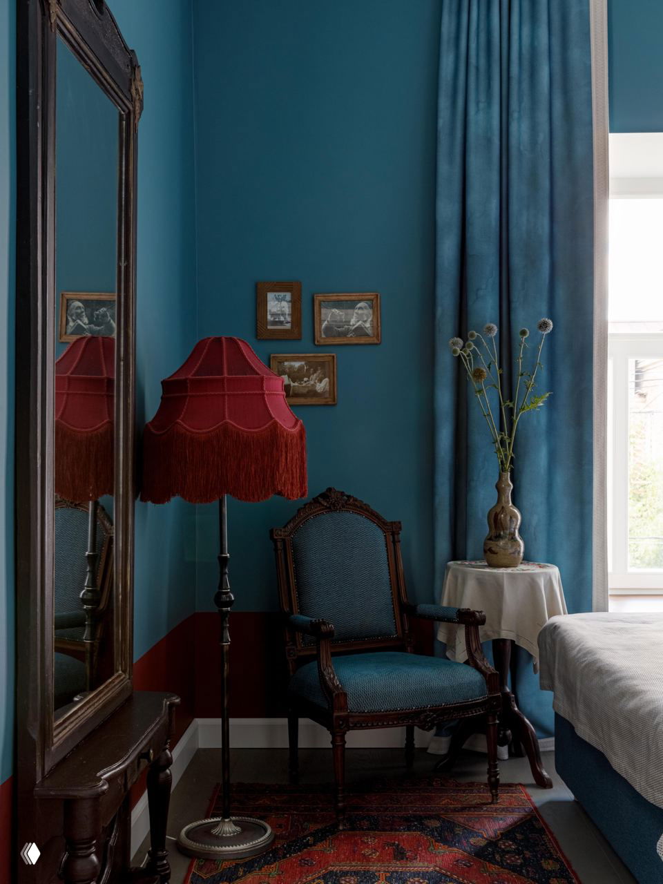 Уютный номер в сине-бирюзовых тонах с высоким зеркалом, торшером с красным абажуром и декоративными элементами в викторианском стиле на фоне плотных штор.