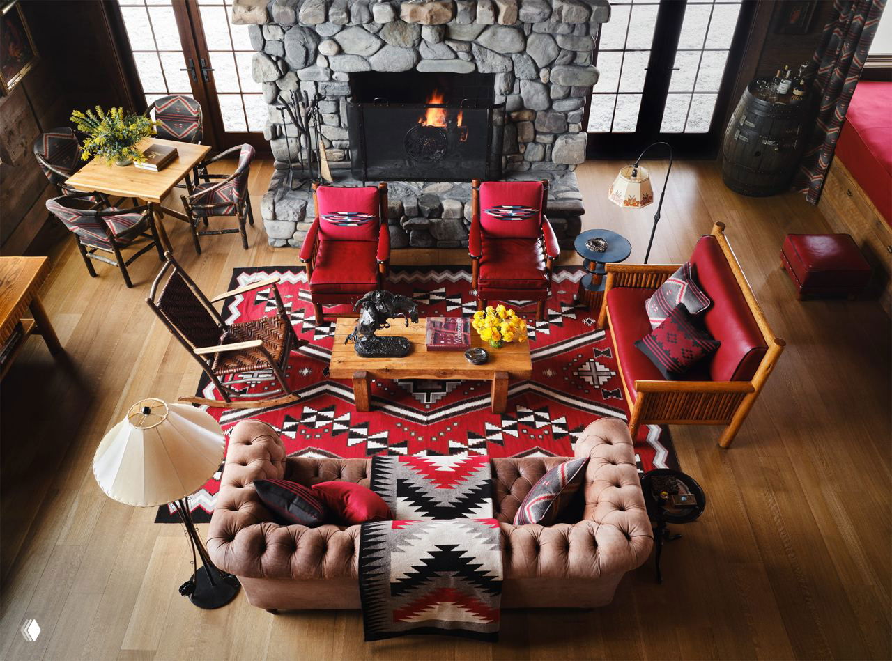 Гостиная с крупным камином из речного камня, групповой расстановкой диванов и кресел, красно-чёрный ковер в навахо-стиле и декоративные аксессуары.