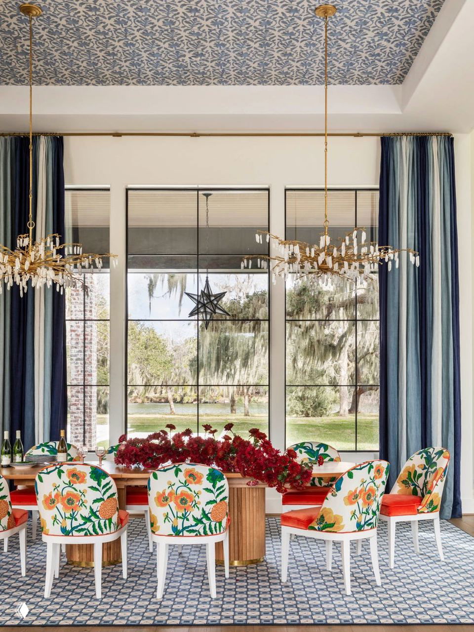 Столовая с панорамными окнами и видом на деревья с мхом: длинный деревянный стол, стулья с цветочным принтом и ковёр с геометрическим узором.