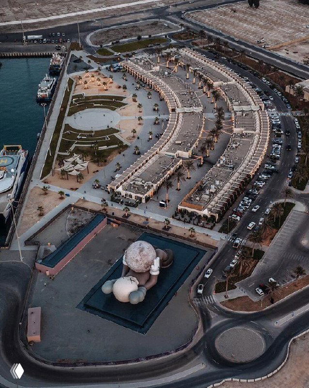 Аэрофотоснимок инсталляции KAWS в порту Мина Зайед: лежащая фигура длиной 32 метра на платформе у воды, окружающая инфраструктура и парковки.