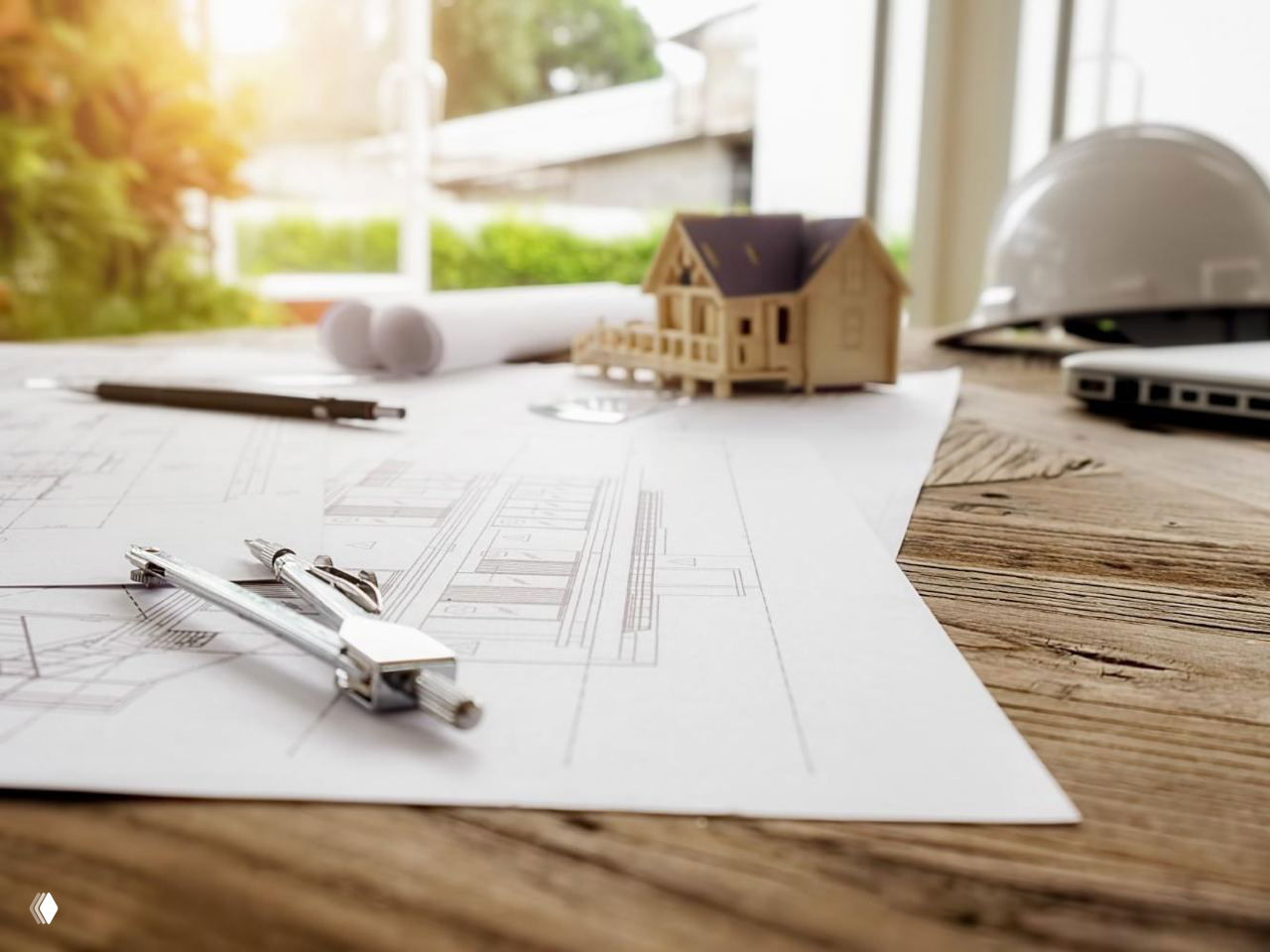 План дома и чертежи на столе в солнечном свете, рядом циркуль и макет деревянного дома, строительная каска на заднем плане.
