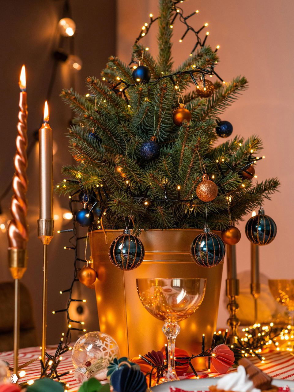 Небольшая настольная живая ёлка в золотом горшке, украшенная миниатюрными гирляндами и тёмно-синими и золотыми шарами на праздничном столе.