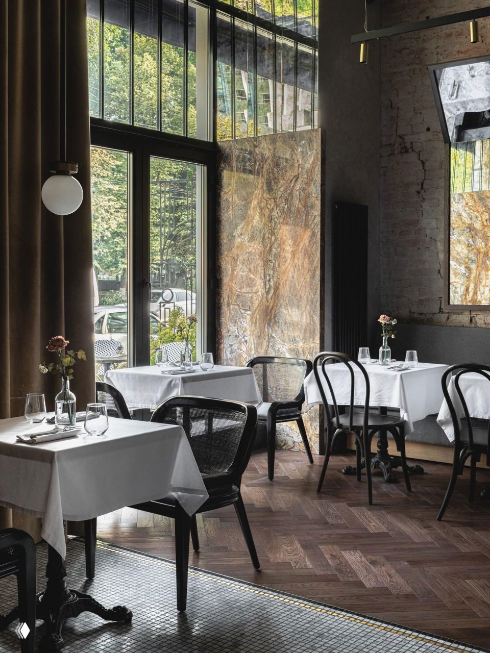 Угол зала у высоких окон: деревянный паркет, столы с белыми скатертями, натуральный камень на стене и зелень за стеклом.