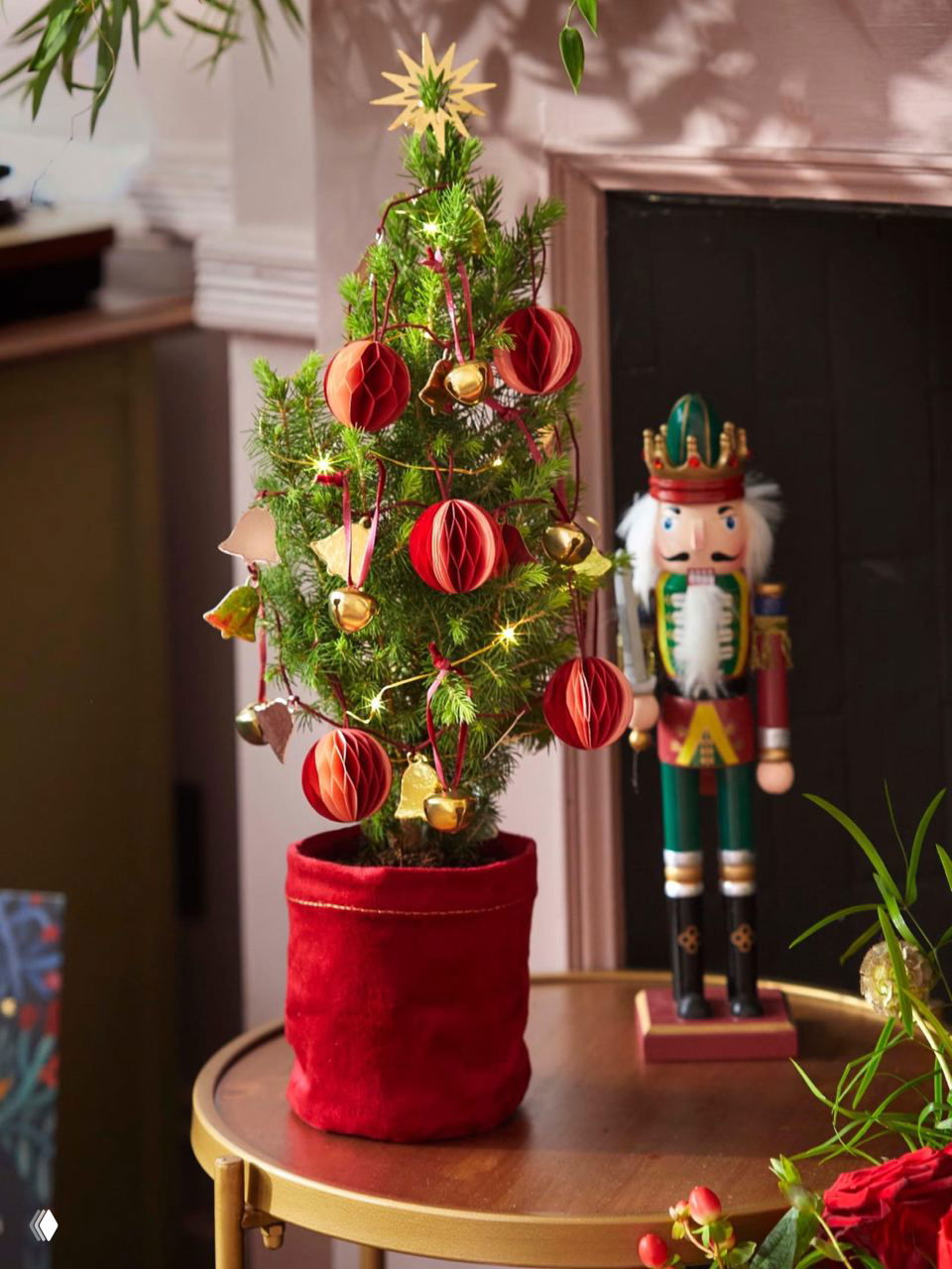 Миниатюрная нарядная ёлка в красном текстильном чехле на столике рядом с игрушкой-щелкунчиком и золотыми гирляндами и шариками.