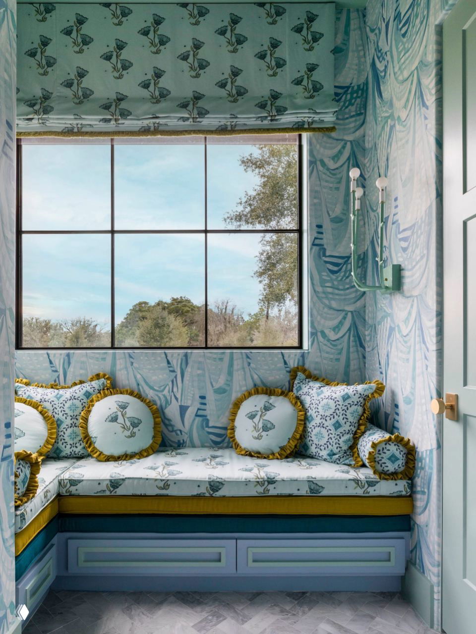 Небольшая уютная ниша у окна: встроенное мягкое сиденье с декоративными подушками, светлые обои с растительным узором и вид на участок ранчо.