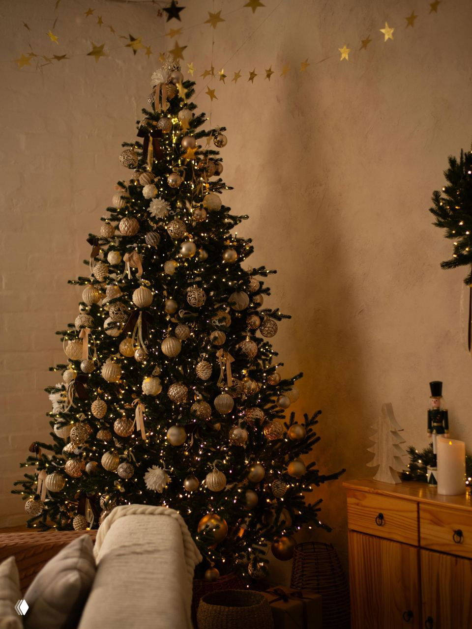 Тёпло освещённая живая ёлка в гостиной с множеством золотых украшений, гирлянд и декоративных элементов в уютной атмосфере вечера.