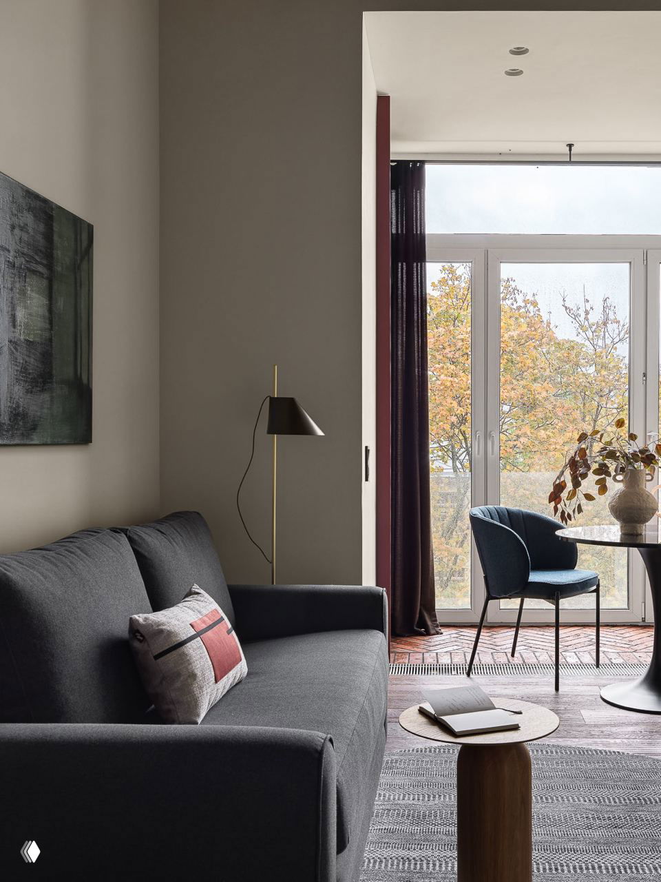 Серая гостиная у большого окна с тёмным диваном, высоким торшером и видом на осенние деревья — сочетание света и глубоких тонов для глубины комнаты.