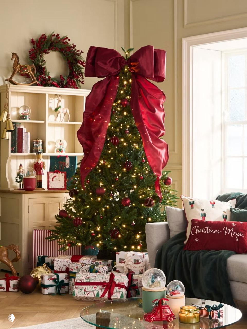 Высокая ёлка с большим бархатным бантом в верхней части, окружённая множеством праздничных подарков и декоративных деталей в гостиной.