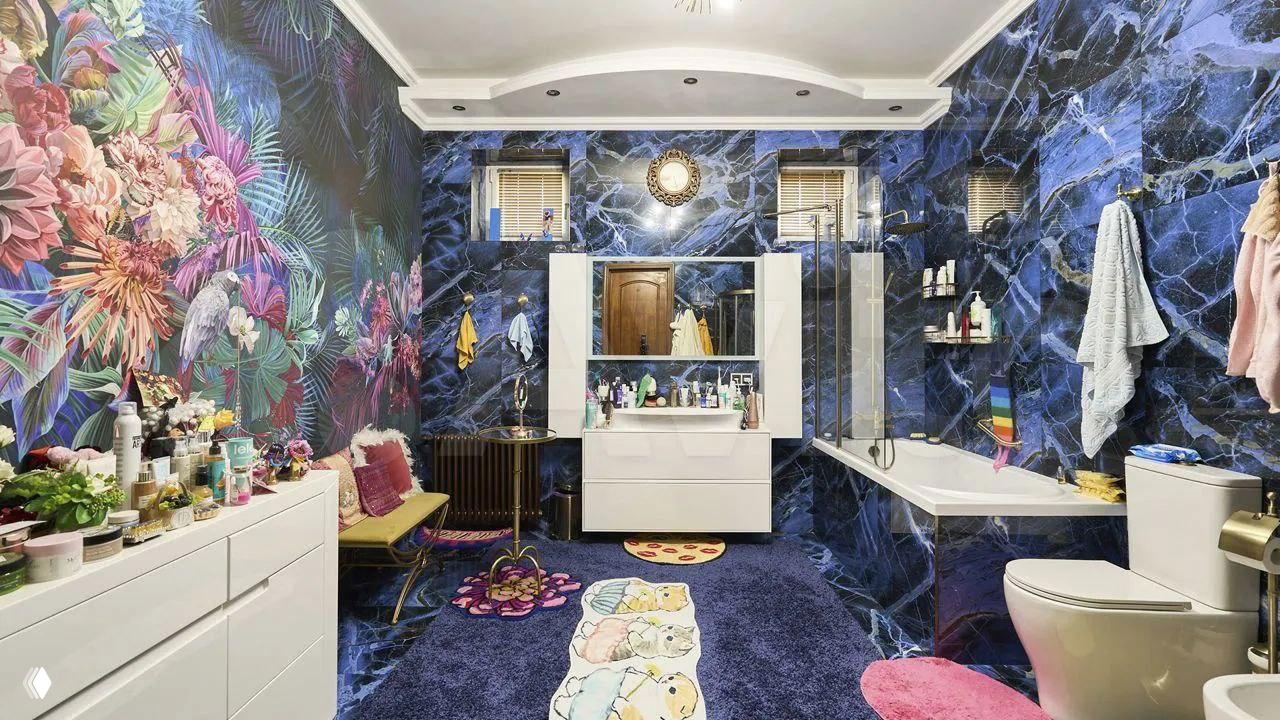 Яркая ванная комната с синими плитками, фотообоями с экзотическими цветами, ковриками с изображением котиков и множеством косметики на тумбах — эклектичный декор.