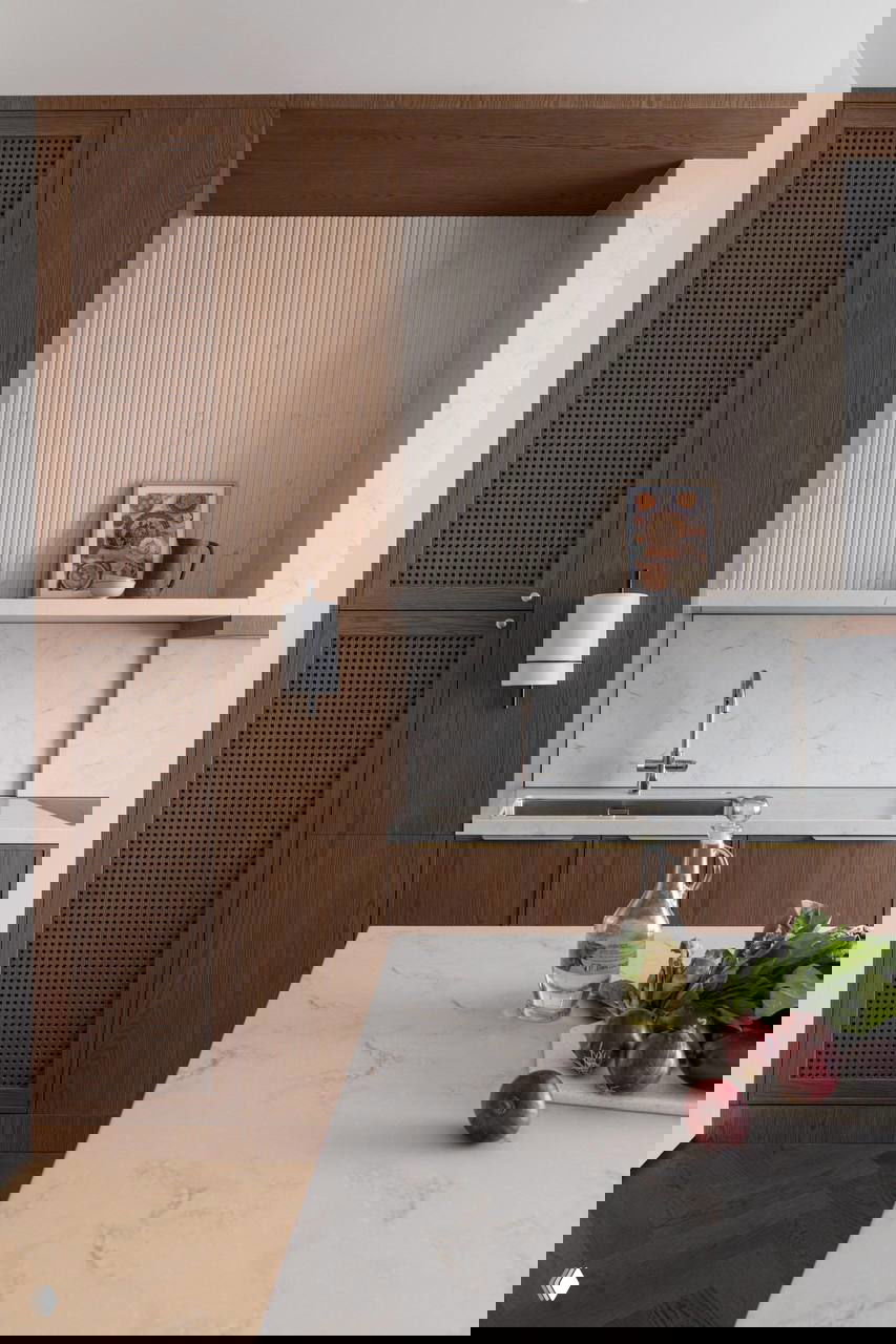 Кухня с каменной столешницей и тёплыми деревянными фасадами в светлом интерьере; на столе масло и красные овощи на переднем плане.