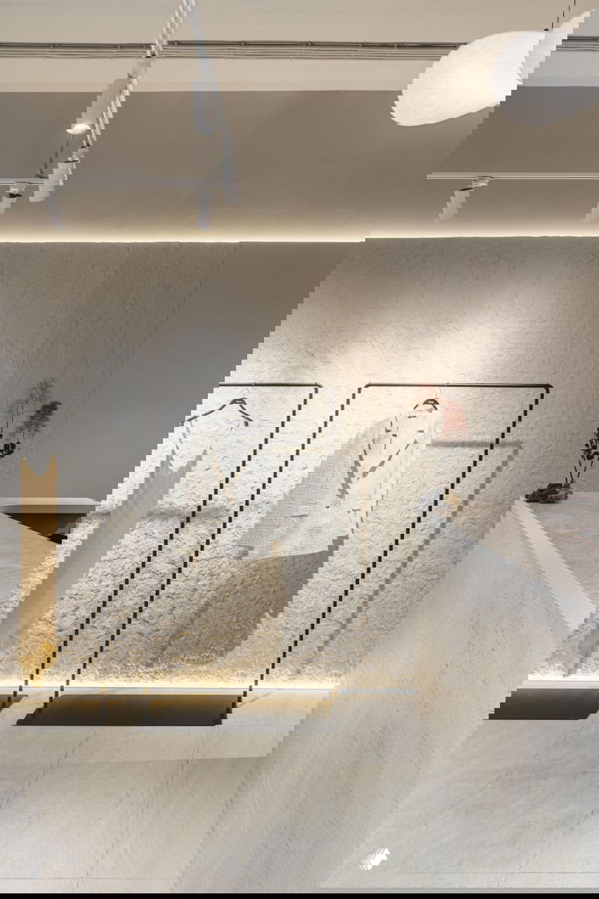 Светлая стена с фактурным скальным рельефом и кассовая зона с отделкой травертином, минималистичные стеллажи и подсветка у пола в бутике Blanco.