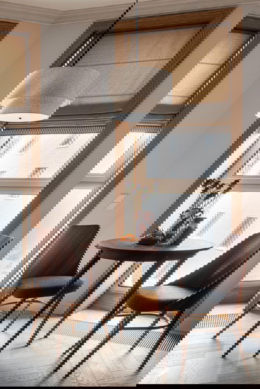 Небольшая обеденная зона у окна: круглый деревянный стол, две стулья из темного дерева и подвесной светильник в нейтральных тонах.