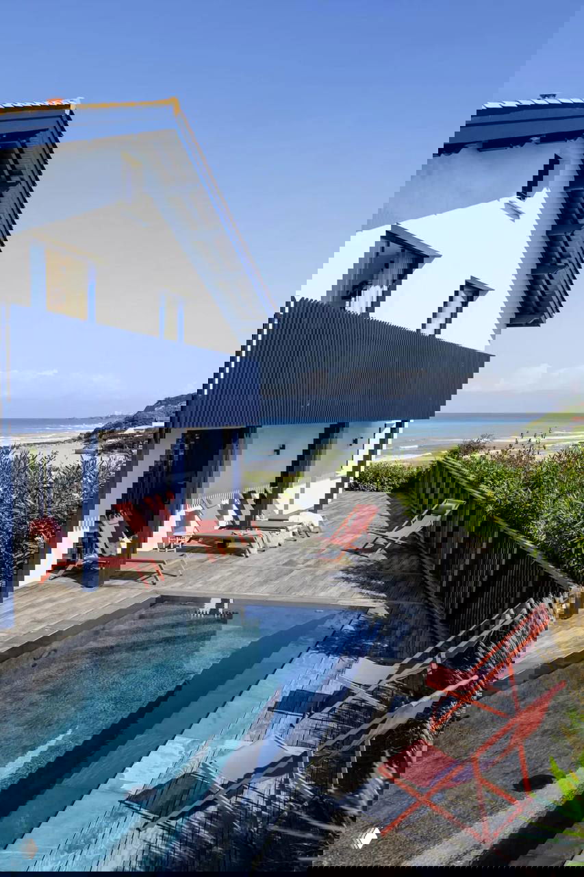 Фасад виллы у пляжа Бидар: деревянная терраса с бассейном, ряд красных шезлонгов и открытый морской горизонт за забором.