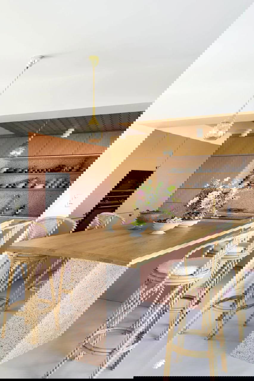 Светлая кухня-столовая: большой деревянный стол-остров, розовые фасады и ротанговые барные стулья в мягкой пастельной гамме.