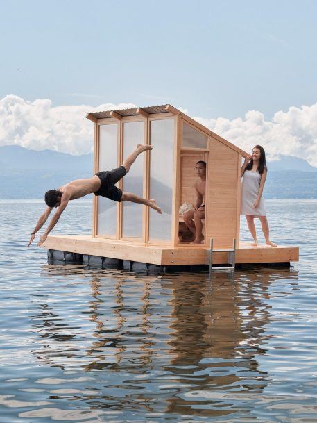 Плавающая финская баня на понтоне: компактная деревянная кабина на воде, люди у входа и прыгают в озеро рядом.