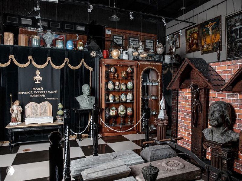 Интерьер Музея мировой погребальной культуры в Новосибирске: витрины с урнами и артефактами, бюсты, траурная символика и музейные ограждения.