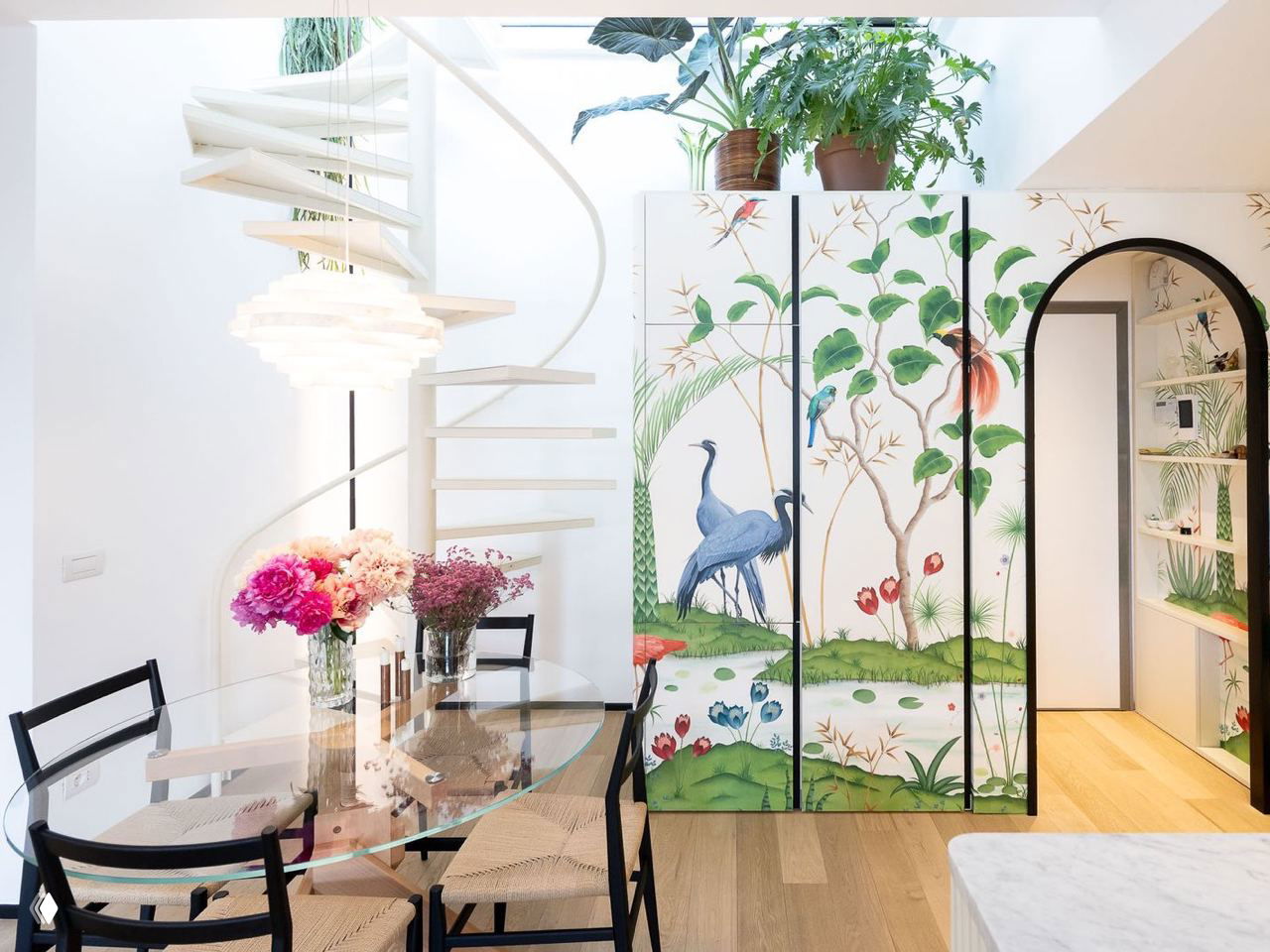 Спиральная белая лестница и декоративные панели с крупным растительным орнаментом, солнечный свет и букеты на столе создают лёгкую атмосферу.