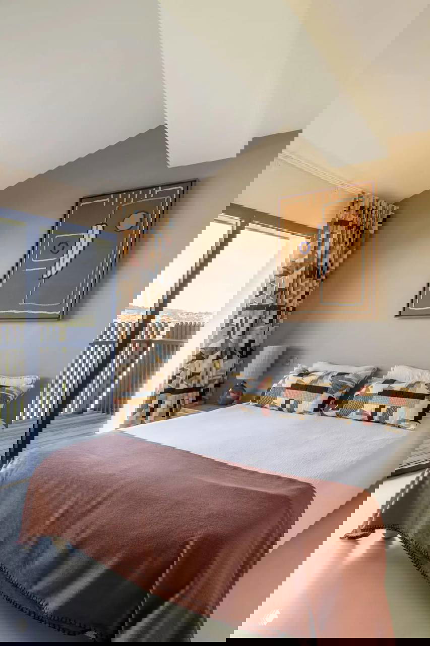 Спальня в нейтральной палитре: большая кровать с терракотовым покрывалом, декоративные подушки и окно-терраса с видом на берег.