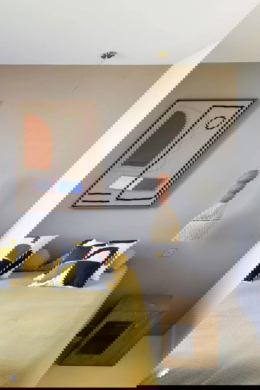 Вторая спальня с охристым покрывалом и висящей кисточковой лампой; мягкие текстуры и абстрактная картина на стене.