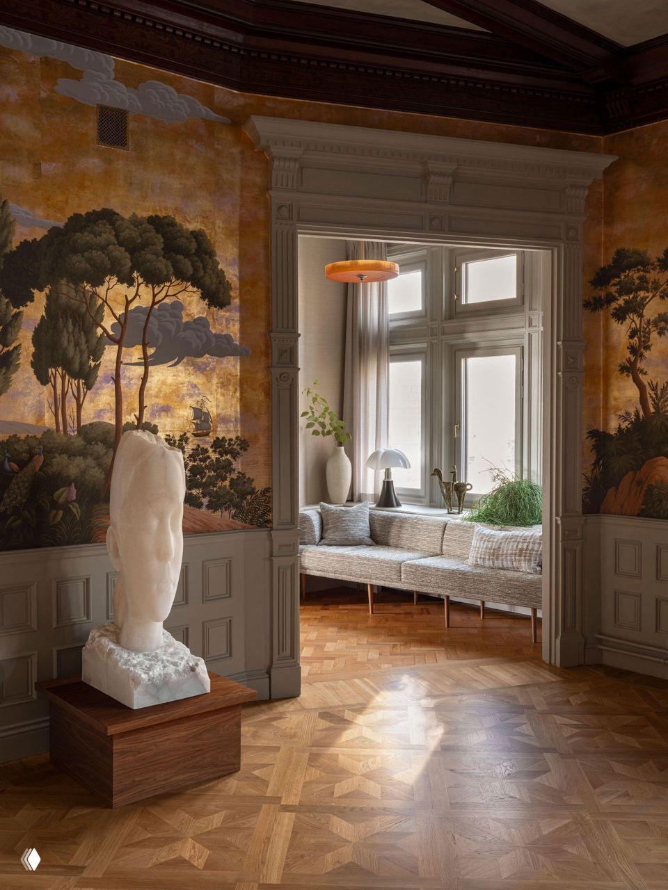 Комната с обширной настенной фреской в золотистых тонах, большим белым скульптурным бюстом на деревянном постаменте и мягким диваном у окна для отдыха и чтения.