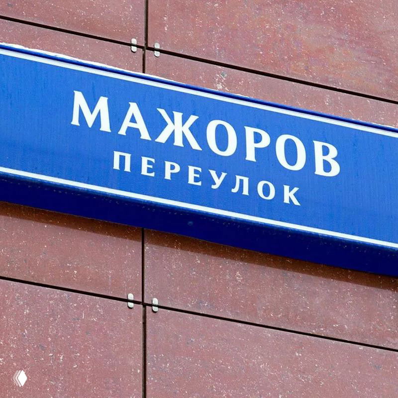 Синяя табличка с белой надписью «МАЖОРОВ ПЕРЕУЛОК» на фасаде кирпичного дома; урбанистический контекст, дорожные знаки и парковка.