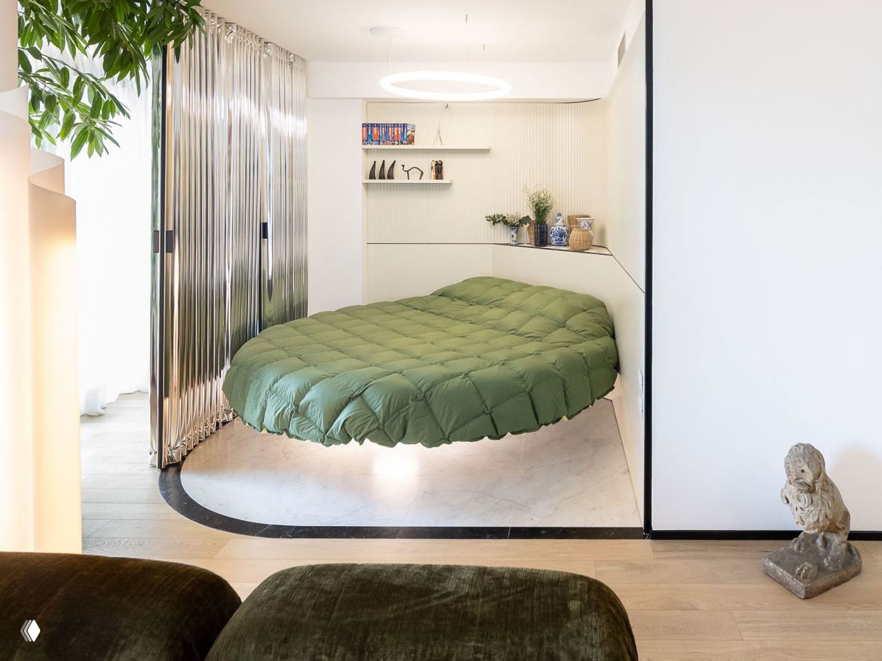 Небольшая спальня за перегородкой: круглая кровать Lago на постаменте, зеленое покрывало и встроенные полки с аккуратными предметами декора.