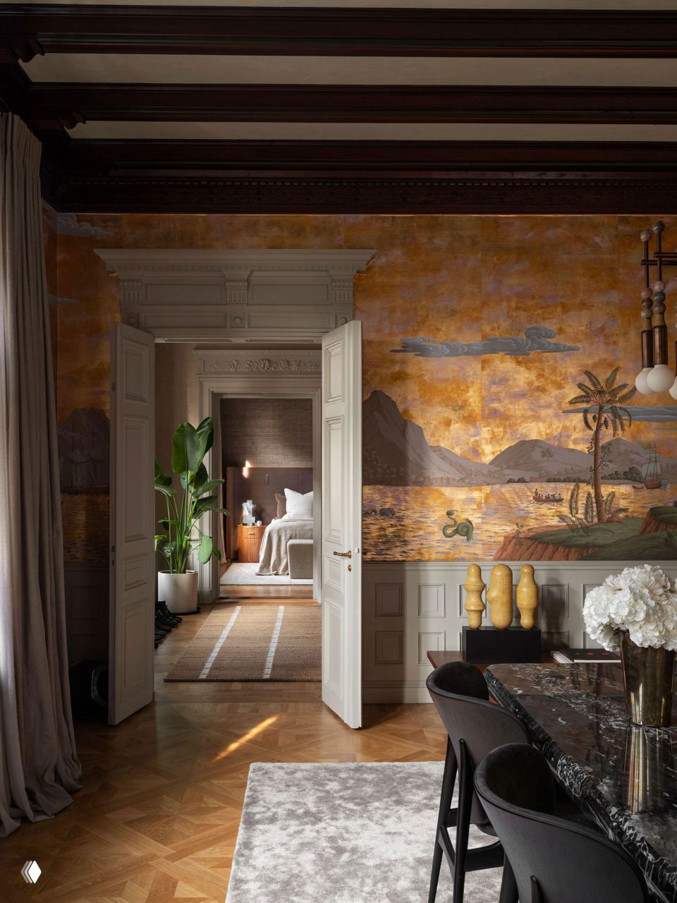 Столовая с декоративной золотистой фреской, деревянными балками на потолке и видом через широкие двери в спальню, где продолжается теплая палитра материалов и текстур.