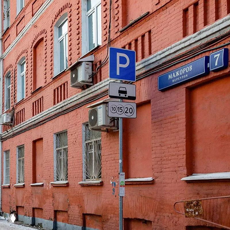 Улица с краснокирпичной застройкой и парковочной разметкой рядом с табличкой переулка; типичный городской фасад и элементы инфраструктуры.