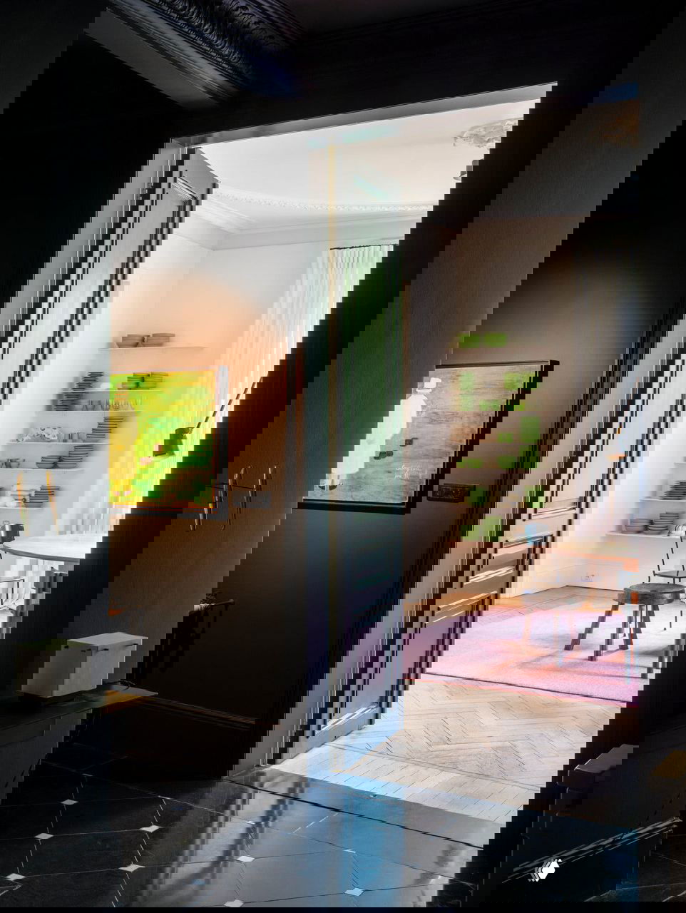 Проход из темной внутренней комнаты в светлую столовую: контраст черной отделки пола и белых стен, видно полки с зелёной посудой и розовый ковёр.