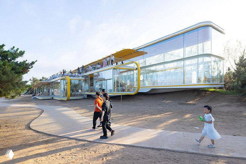 Панорамный вид на стеклянное кафе Chiringuito от Selgascano на пляже Жичжао: прозрачные фасады, песчаная береговая линия, деревья и морской горизонт.