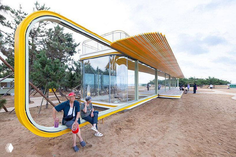 Фрагмент здания с вытянутыми стеклянными объёмами над пляжем: конструкция подчёркивает игру света и цвета, рядом видны отдыхающие и песок.