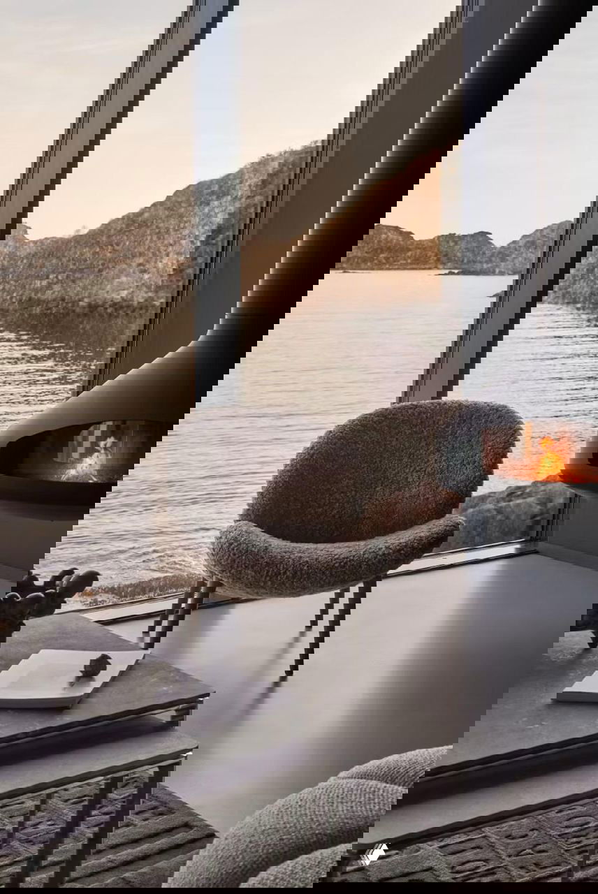 Уютный интерьер с мягким креслом у панорамного окна и подвесным камином, с видом на море и скалистые острова Лофотен.