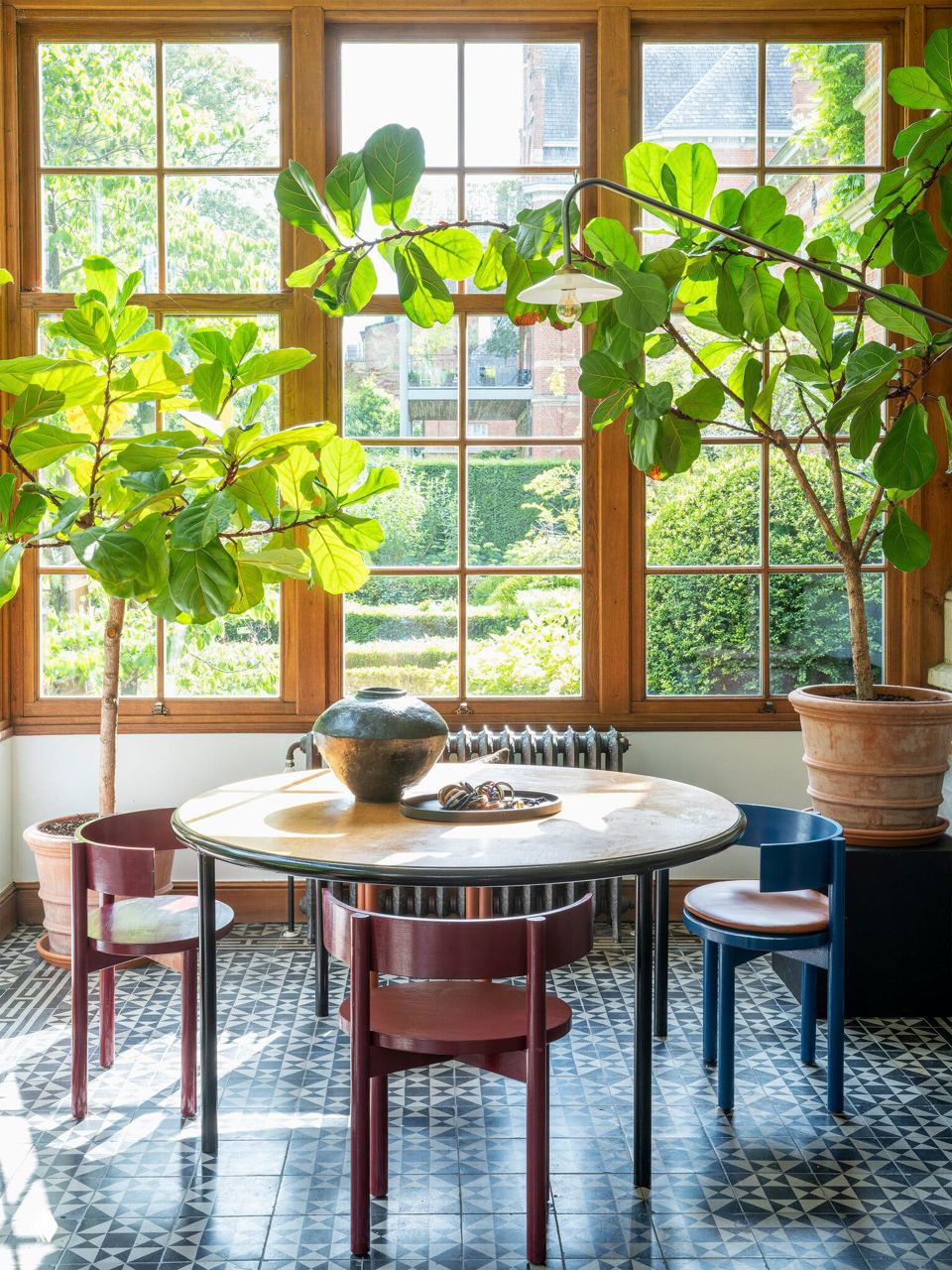 Окно с большим остеклением, видом на сад и двумя фикусами в горшках у круглого обеденного стола на плиточном полу с орнаментом.