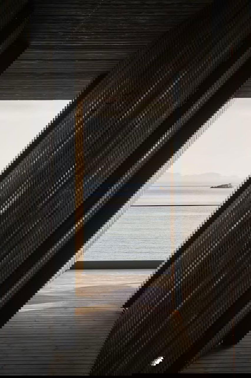 Длинный деревянный коридор между тёмными фасадами хижин, открывающий вид на спокойную морскую поверхность и дальние острова.