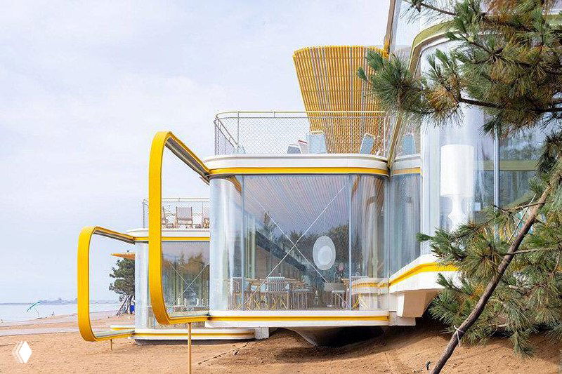 Боковой ракурс кафе Chiringuito: три параллельных стеклянных прямоугольника, смещённые по высоте, на фоне прибрежной растительности и моря.