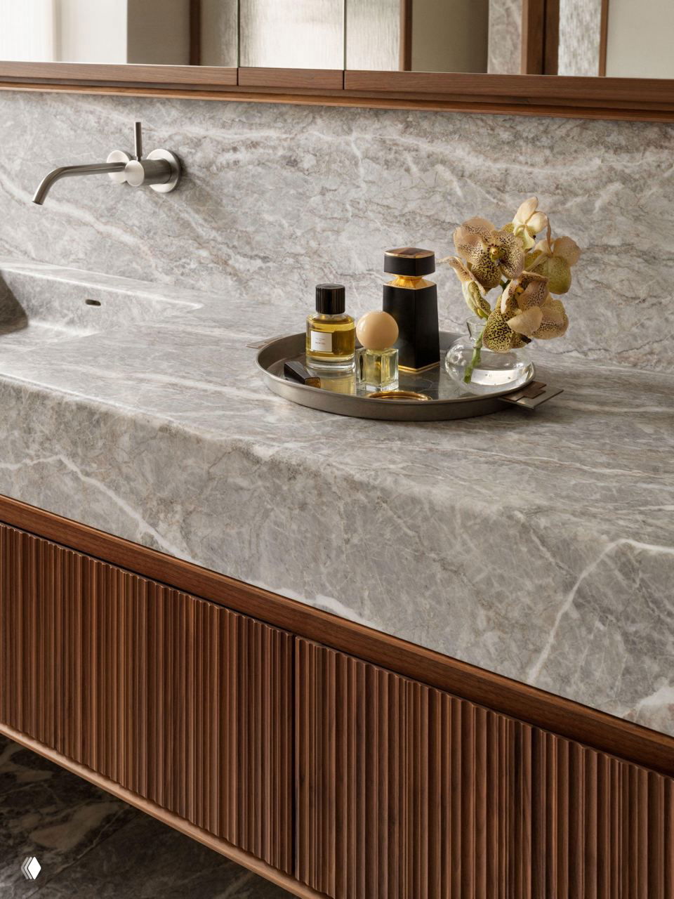 Крупный план мраморной столешницы с серебристой врезной раковиной, декоративным подносом с косметикой и аккуратной цветочной композицией, подчёркивающей материалы интерьера.