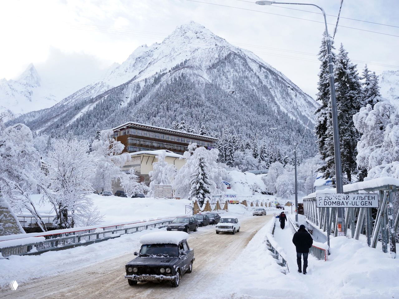 Заснеженная горная дорога на Кавказе с автомобилями и пешеходом, вид на поселок и высокие снежные вершины зимой.