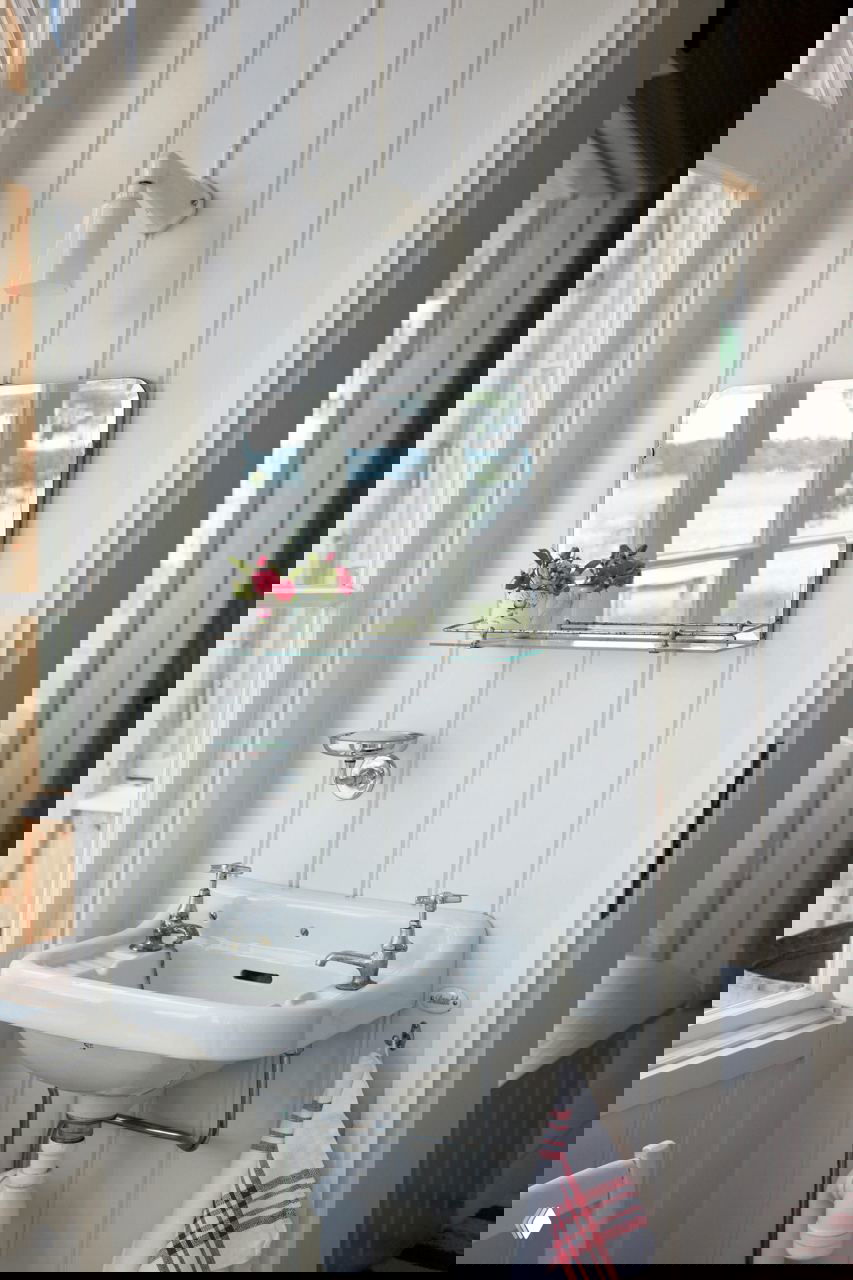 Небольшая зона с настенным зеркалом и фарфоровой раковиной в ретро-стиле на фоне белой вагонки и окна с видом на воду — аккуратный хозяйственный уголок.