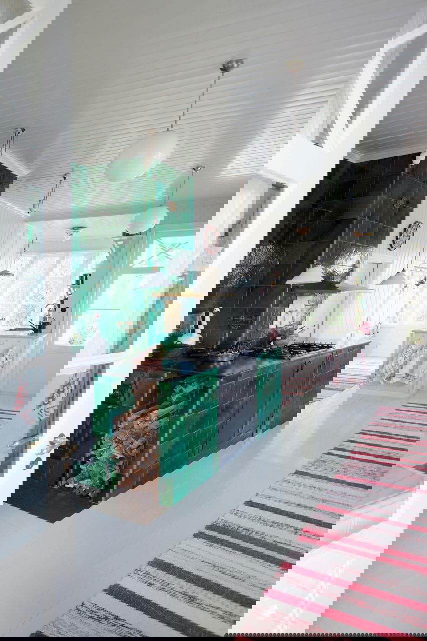 Кухня со стеной из зелёной глазурованной плитки, чёрной столешницей, местом для дров и полосатыми ковровыми дорожками на белом полу в скандинавском стиле.