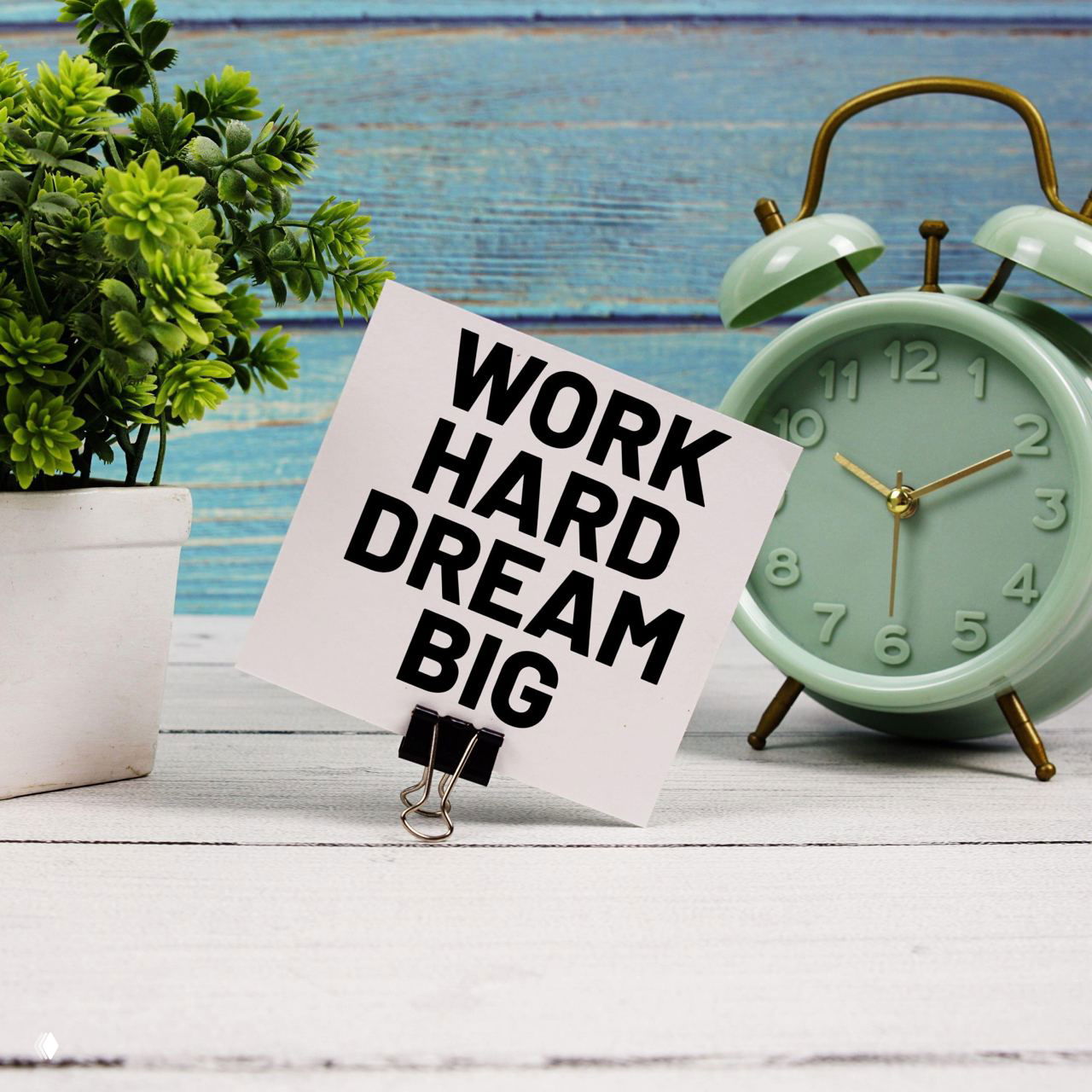 Записка с мотивационной фразой «WORK HARD DREAM BIG» рядом с будильником и маленьким растением — пример трендового мотивационного декора, заметного в интерьере.