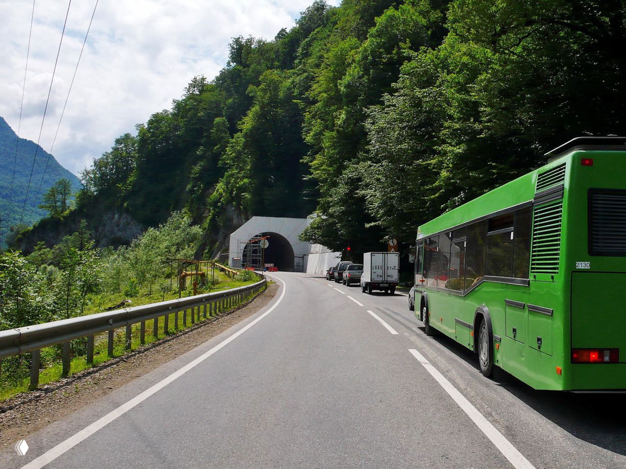 Асфальтированная горная трасса у входа в туннель, стоящие автомобили и зелёный автобус на обочине, густой лес на склонах.