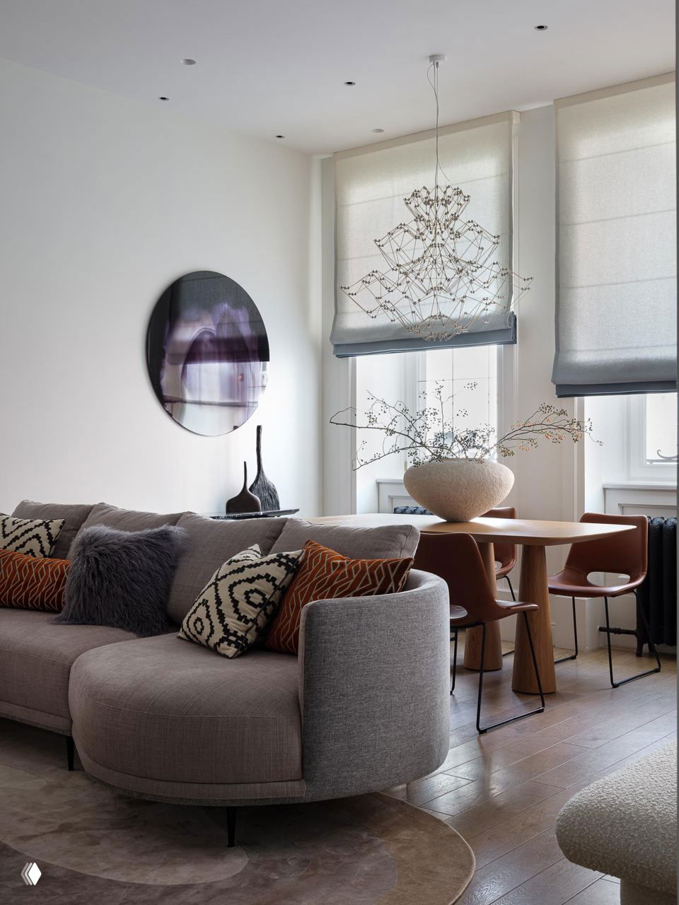 Гостиная с двумя окнами: округлый серый диван, столовая зона у окна и декоративная подвесная люстра в светлом интерьере.
