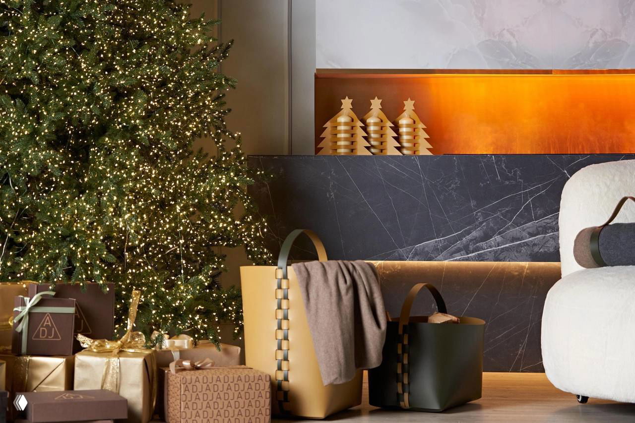 Праздничная интерьерная сцена: украшенная ёлка, подарочные коробки с логотипом и кожаные плетёные сумки‑корзины ADJ на фоне каминной зоны.