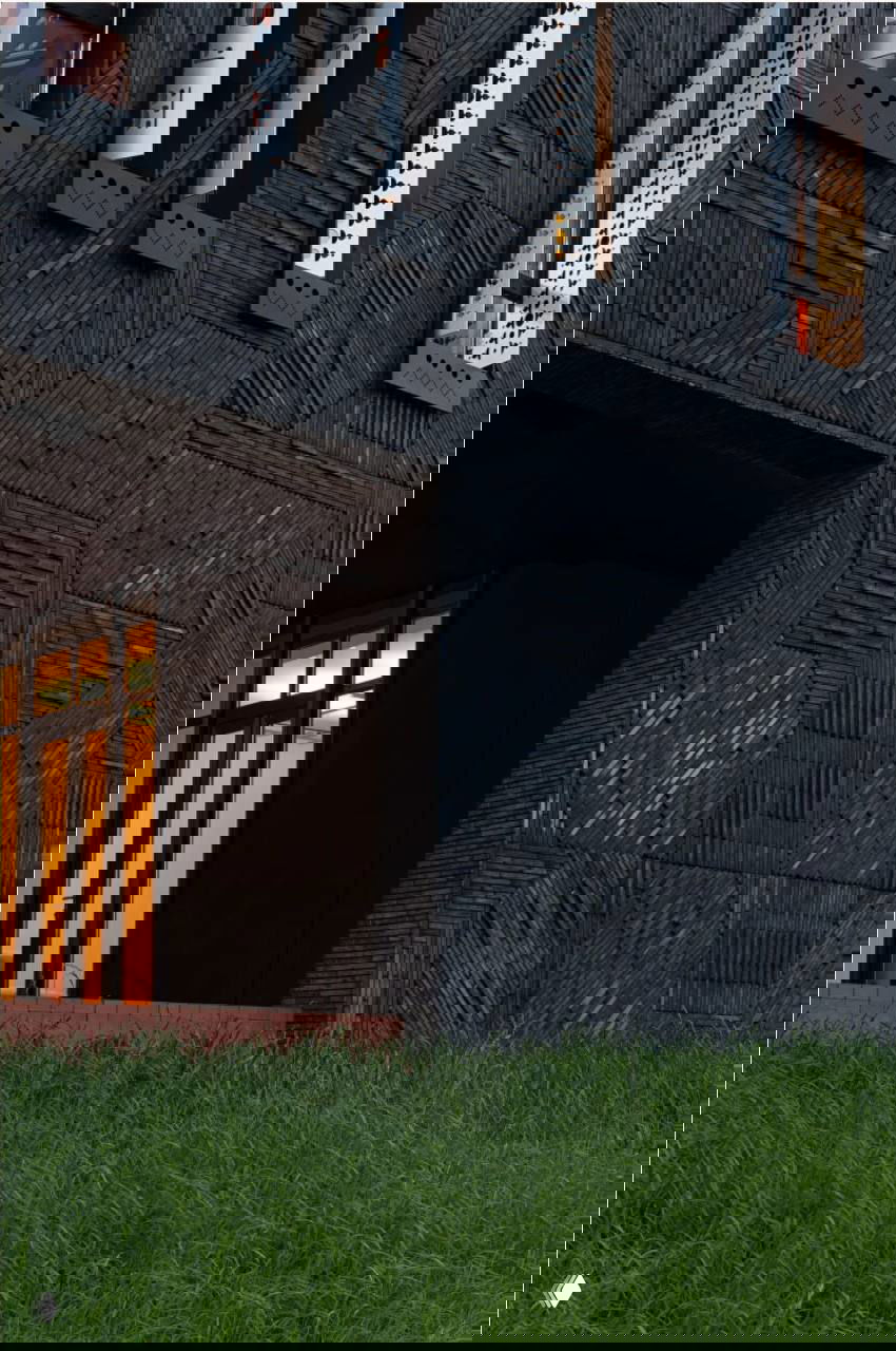 Черный фасад гостевого дома с выразительной текстурой облицовки и большими вертикальными окнами, вид на вход и зеленый покров травы перед зданием.